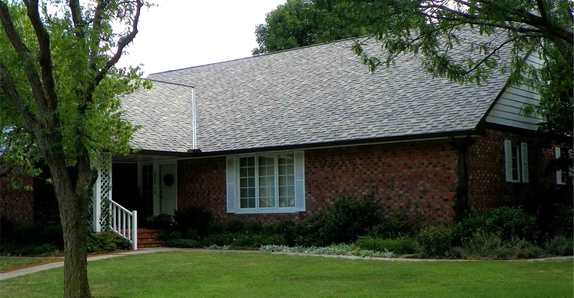 A red brick, single-story house with a grey shingled roof, a white-framed bay window, and a small front porch.