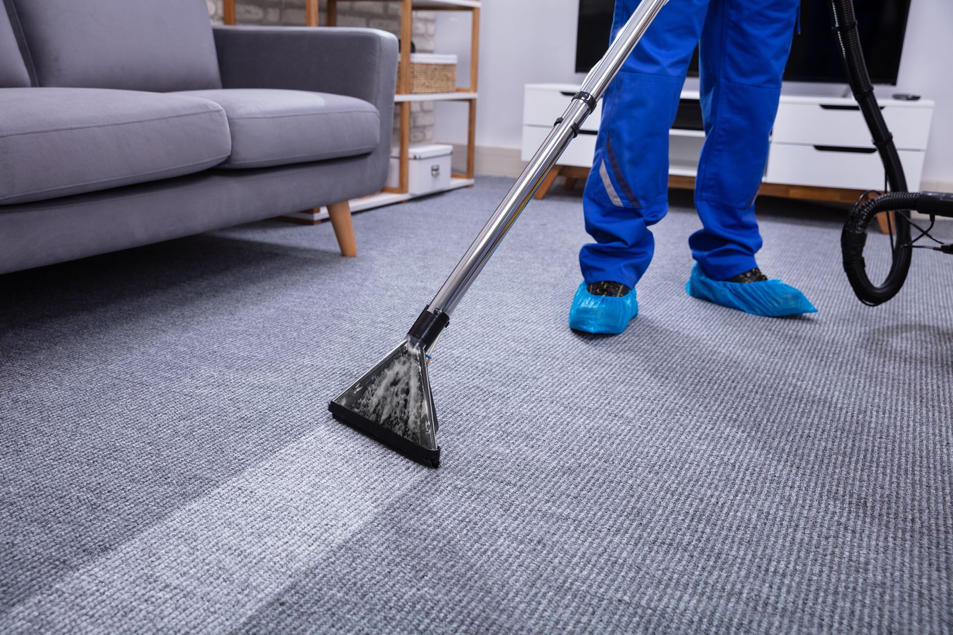 Person in blue coveralls cleans carpet with a steam cleaner in a living room.