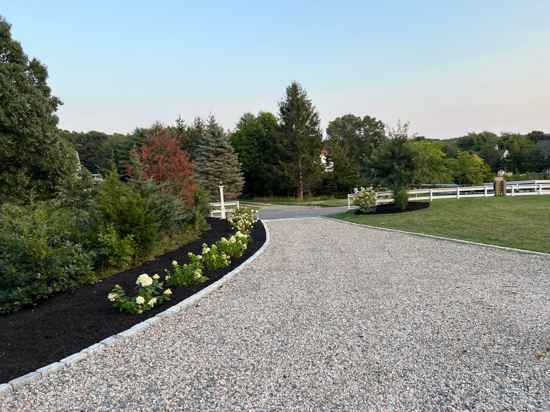 A gravel driveway leading to a lush green field with a white fence