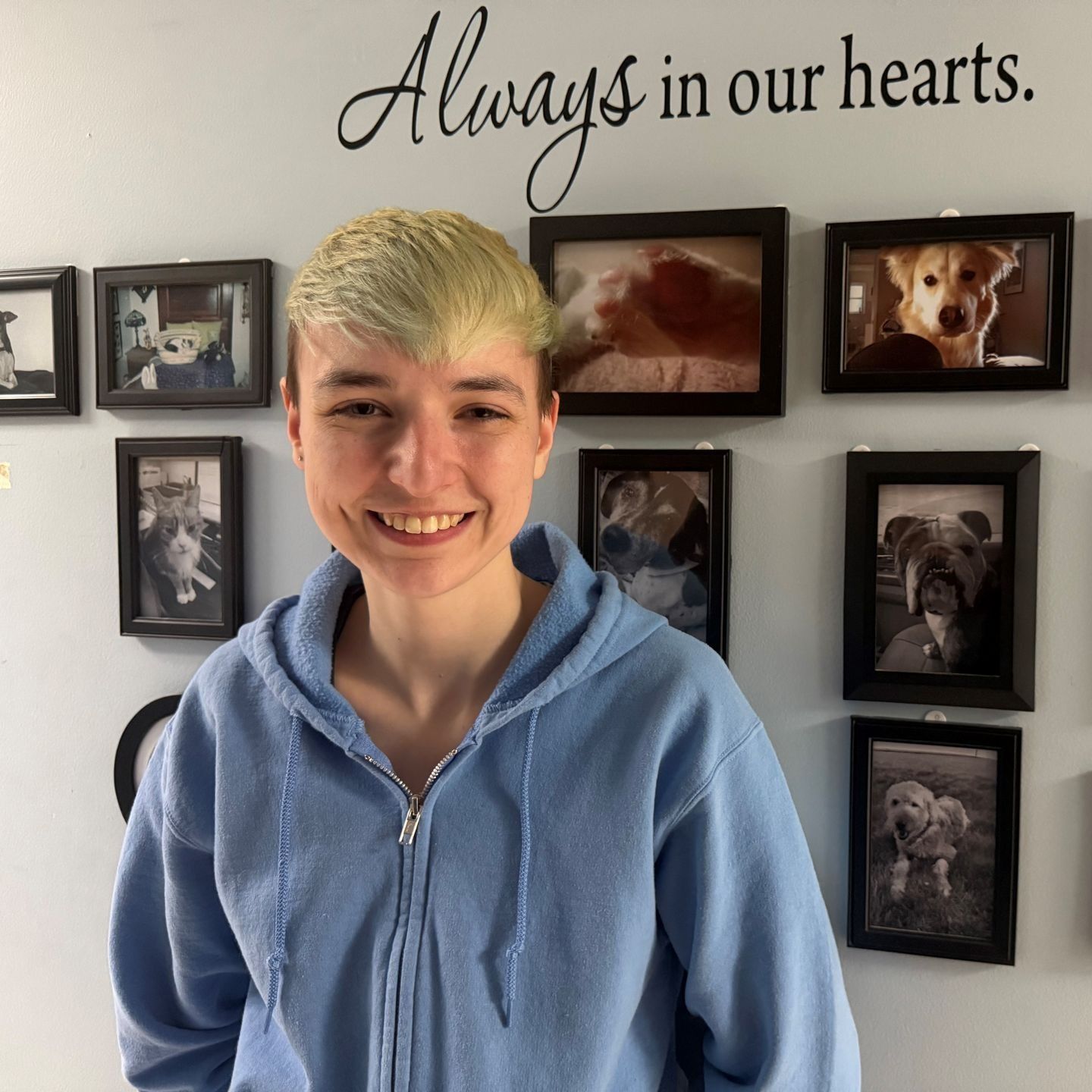 Person in blue hoodie smiles in front of a wall with framed pet photos and
