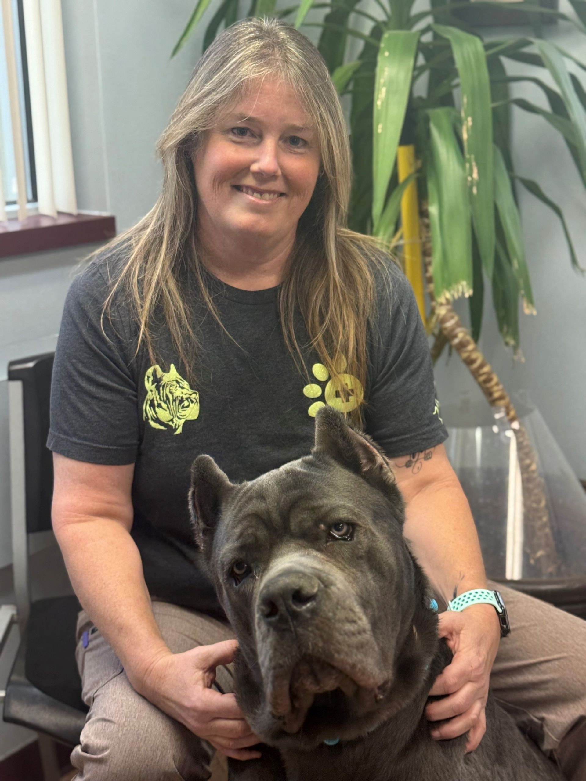 Woman seated with a large gray dog; both are smiling. Indoor setting with a plant.