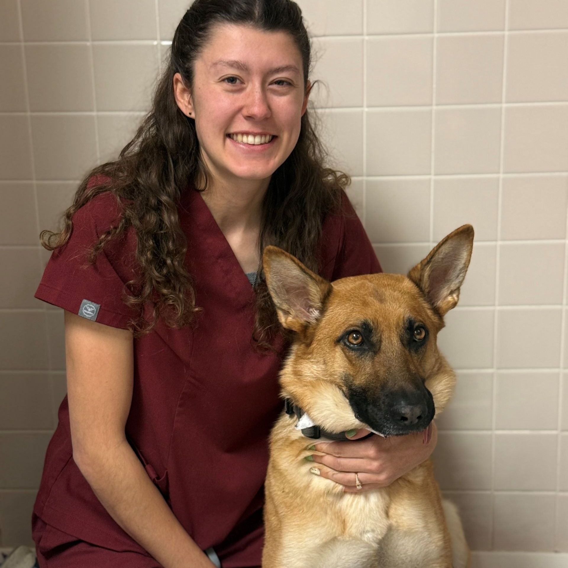Woman in maroon scrubs with a German Shepherd dog; both smiling in a clinic setting.