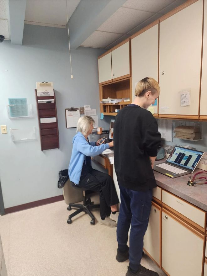 Two people in a room with a desk and cabinets; one uses a computer, the other stands by a laptop.