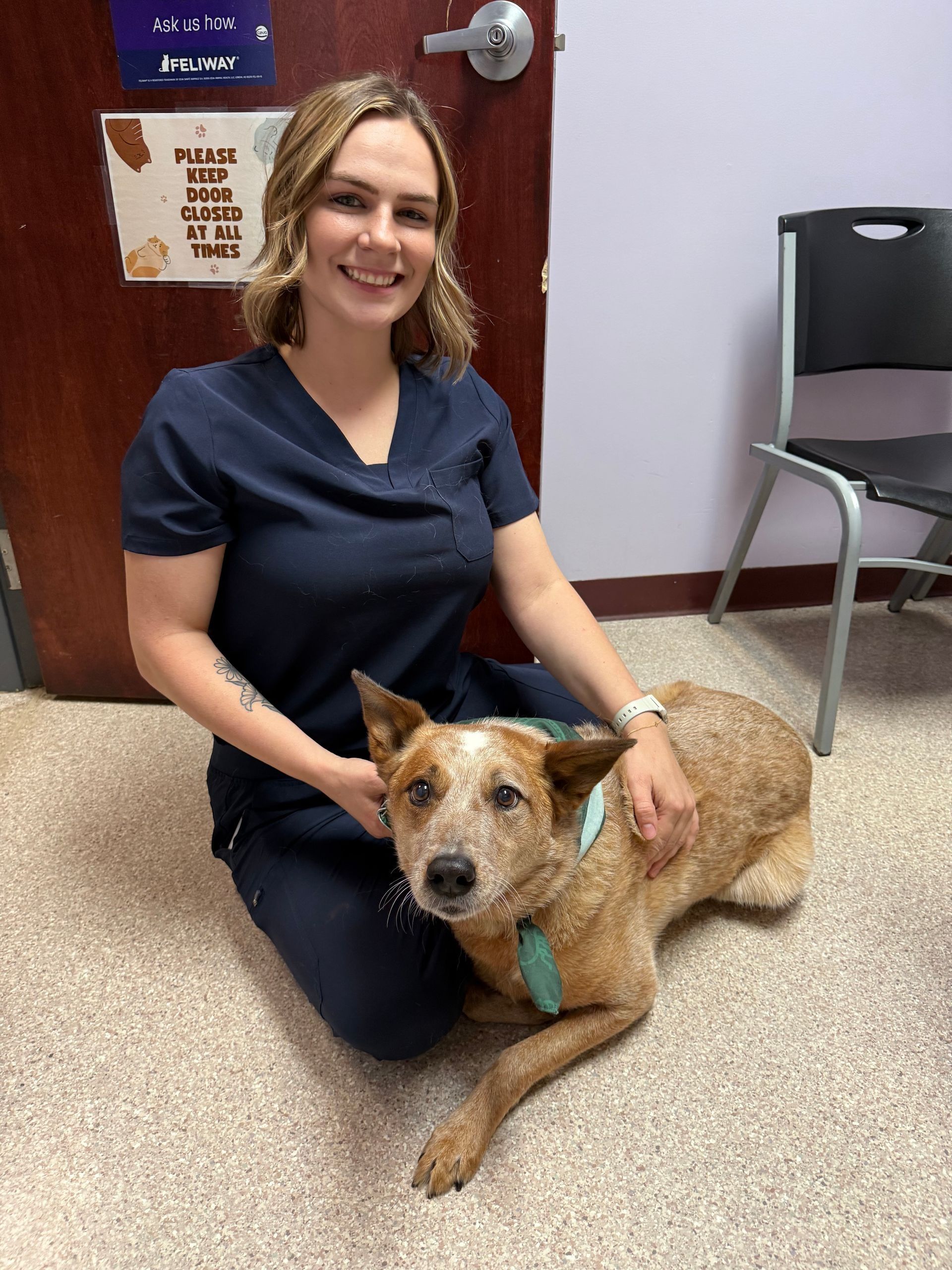 Woman in scrubs kneels with a red and tan dog indoors; both smile.