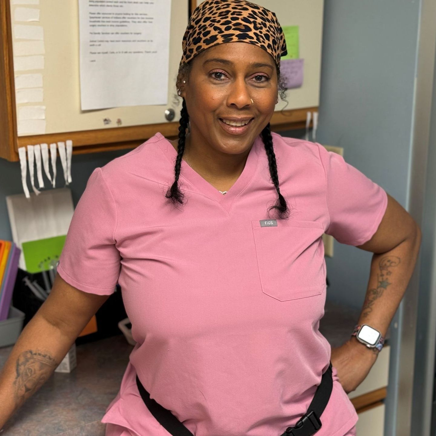 Person wearing pink scrubs and a leopard print headscarf smiling, standing near a desk.