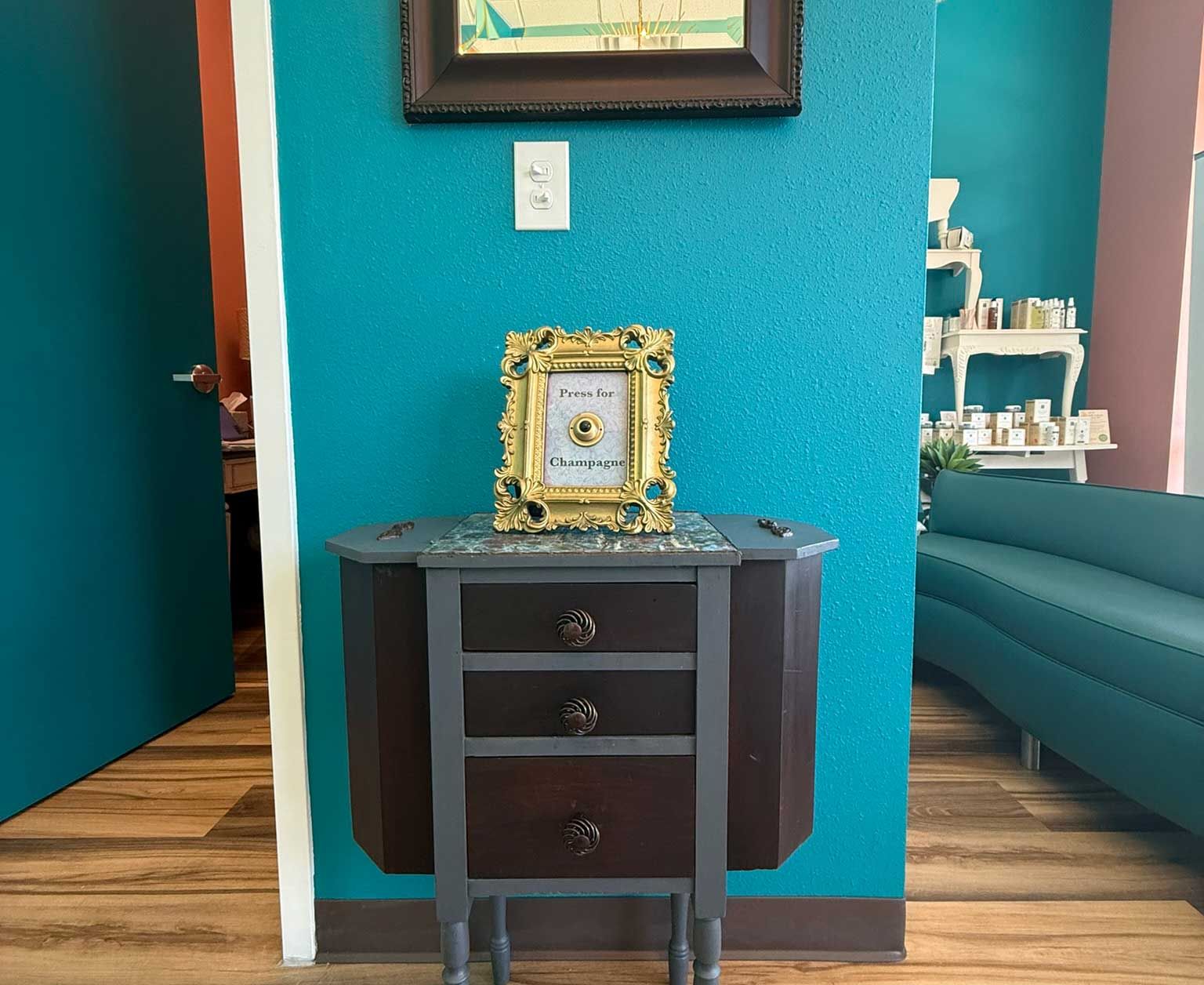 An antique cabinet with a framed item sits against a teal wall. A doorway and sofa are visible.