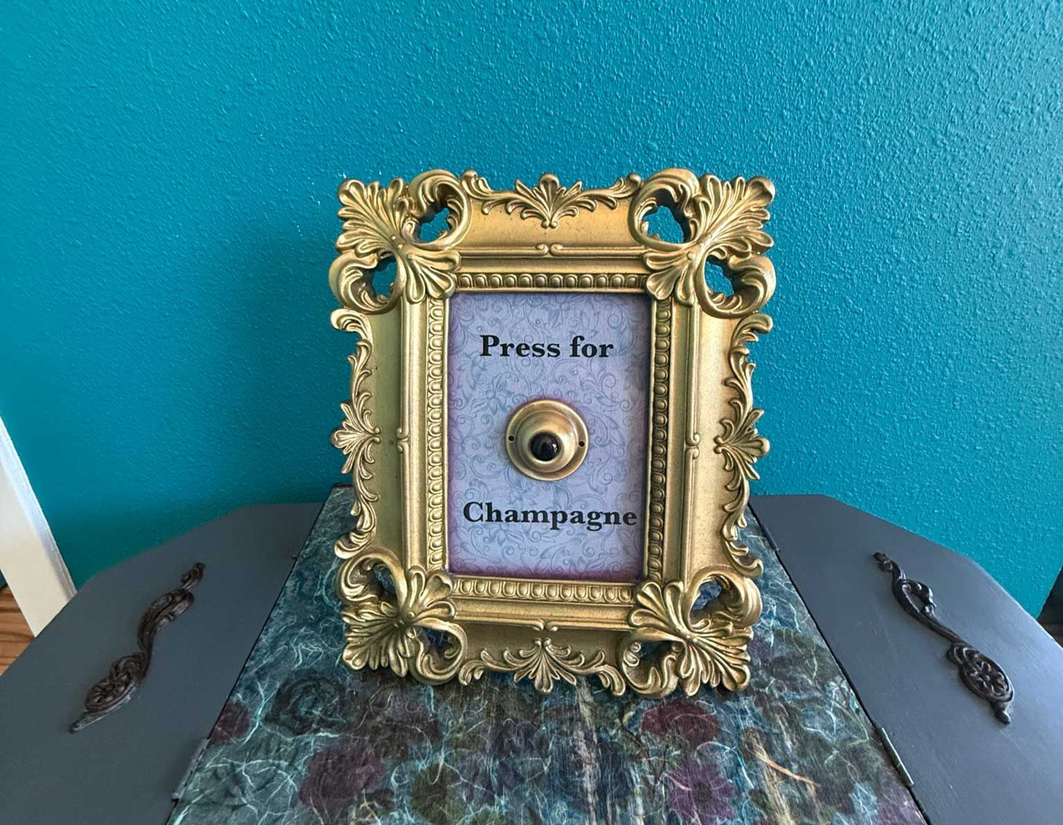 Gold framed button with text, "Press for Champagne," on a table against a blue wall.