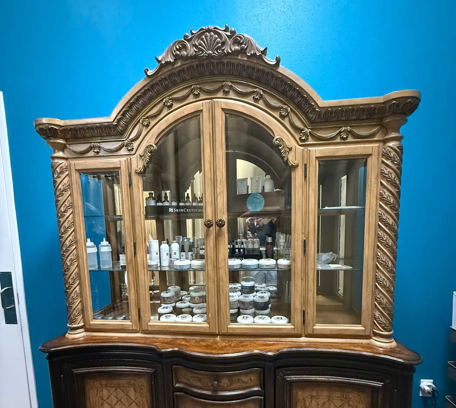 Wooden display cabinet with glass doors filled with beauty products against a blue wall.