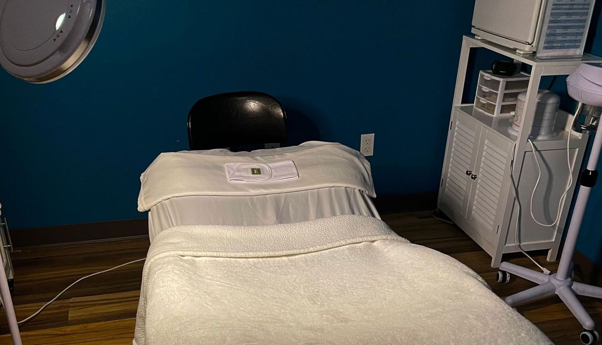 A neatly prepared spa or treatment room with a massage bed covered in white linens, a folded towel on top, and a blue wall background with skincare equipment and shelves nearby.
