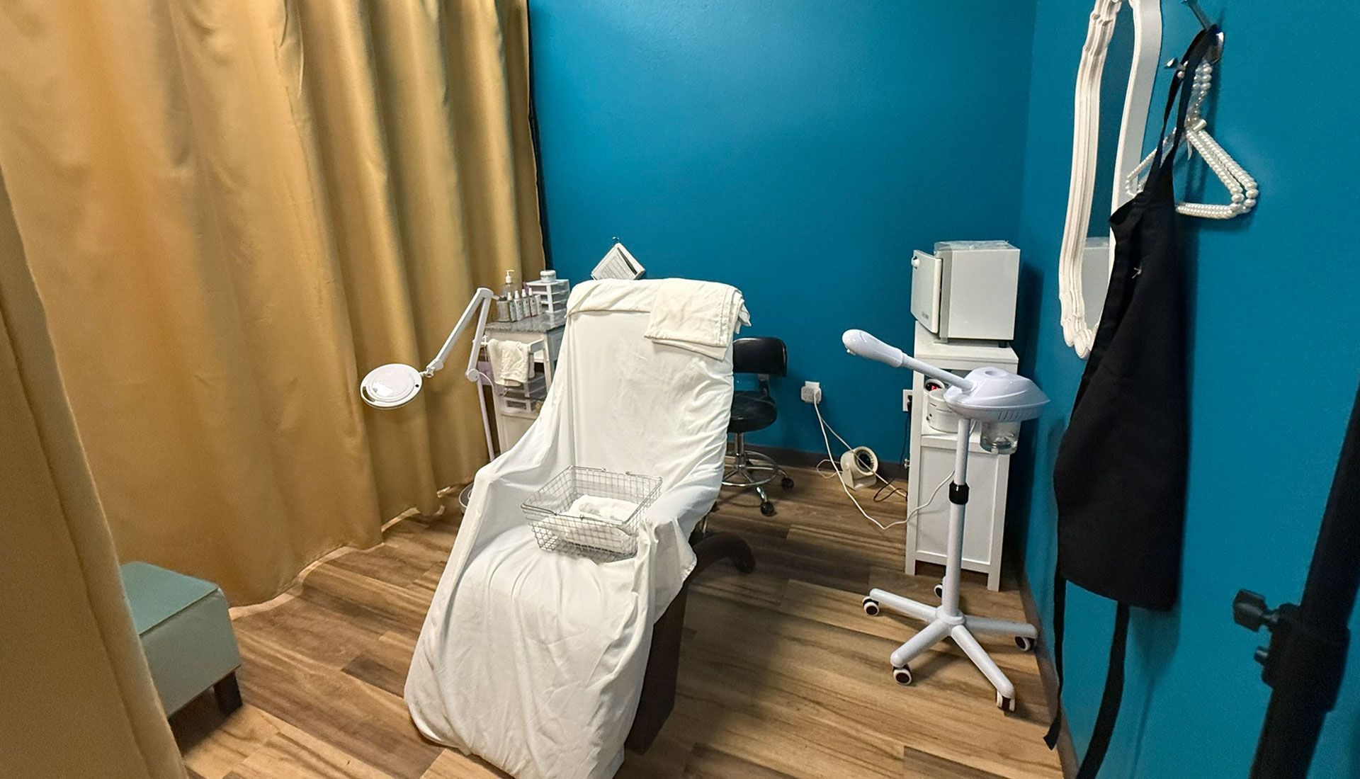 A facial room with a white chair, teal walls, and beige curtain. Medical equipment and a black apron are visible.