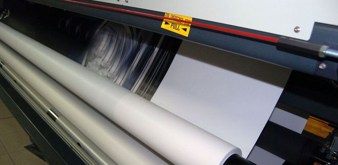 Wide-format printer feeding white paper rolls with ink, printing a banner inside a workshop