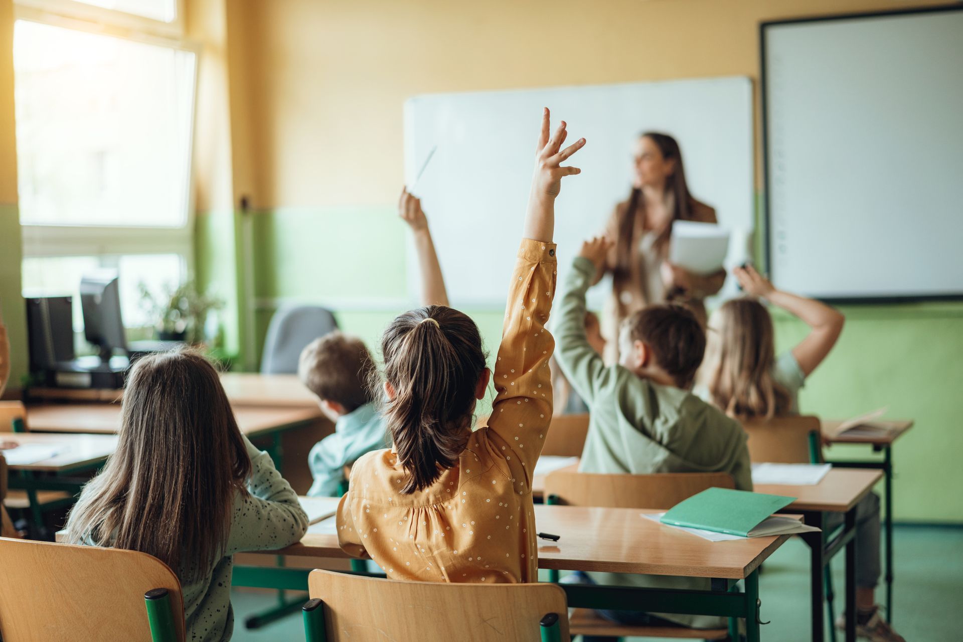 Students raise hands in a classroom, teacher in the background.