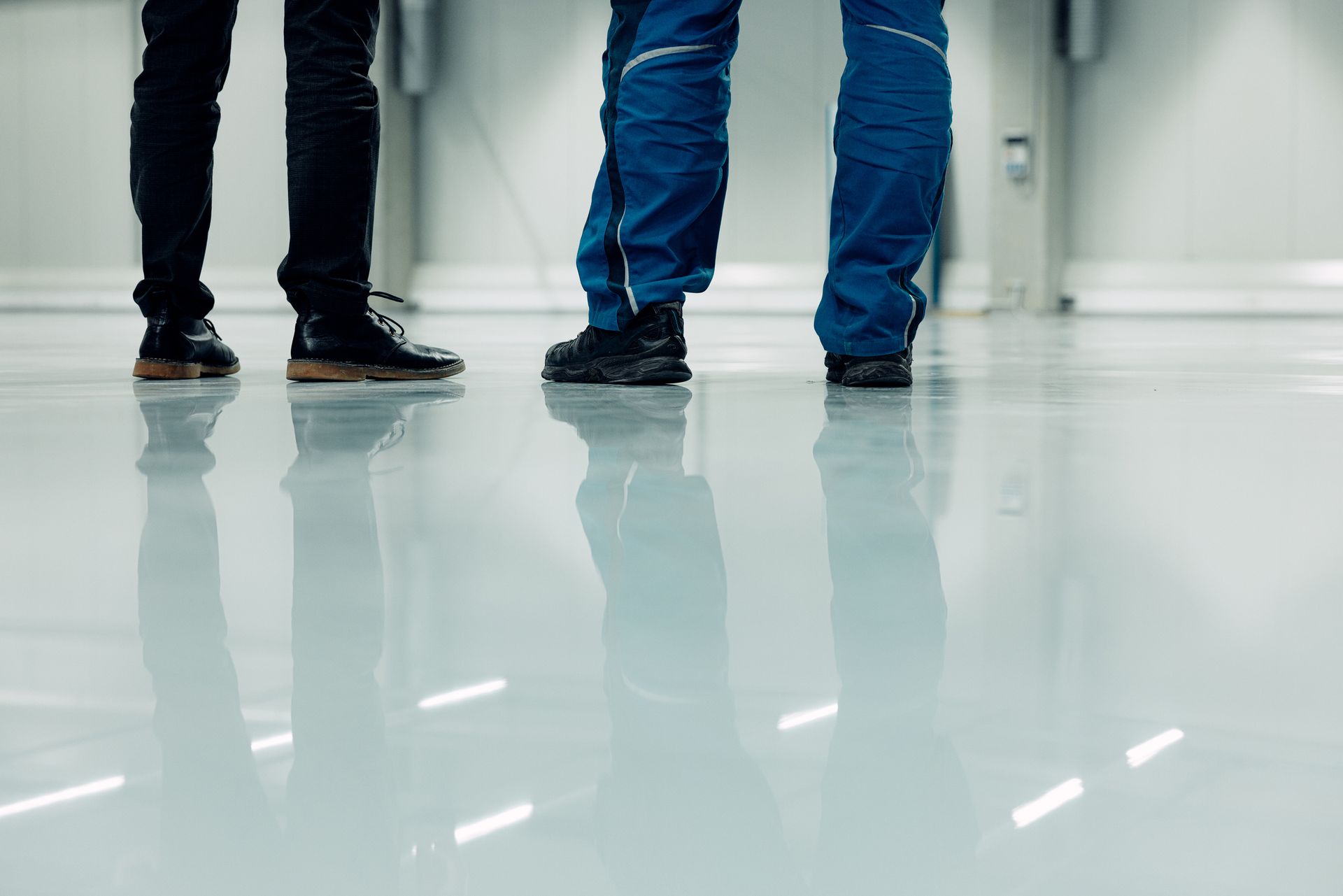 Three people's legs and feet, two wearing dark pants, one in blue, standing on a reflective, shiny floor.