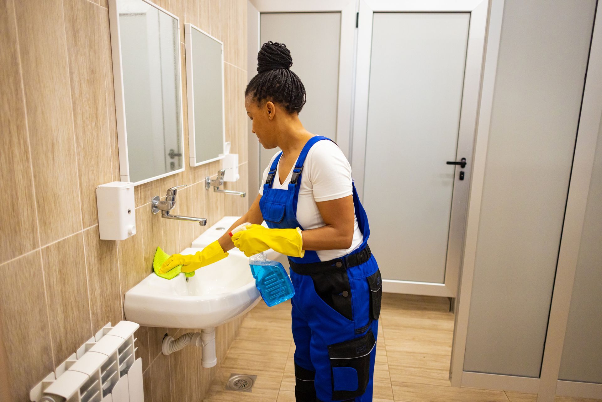 Person in blue overalls and yellow gloves cleaning a bathroom sink.