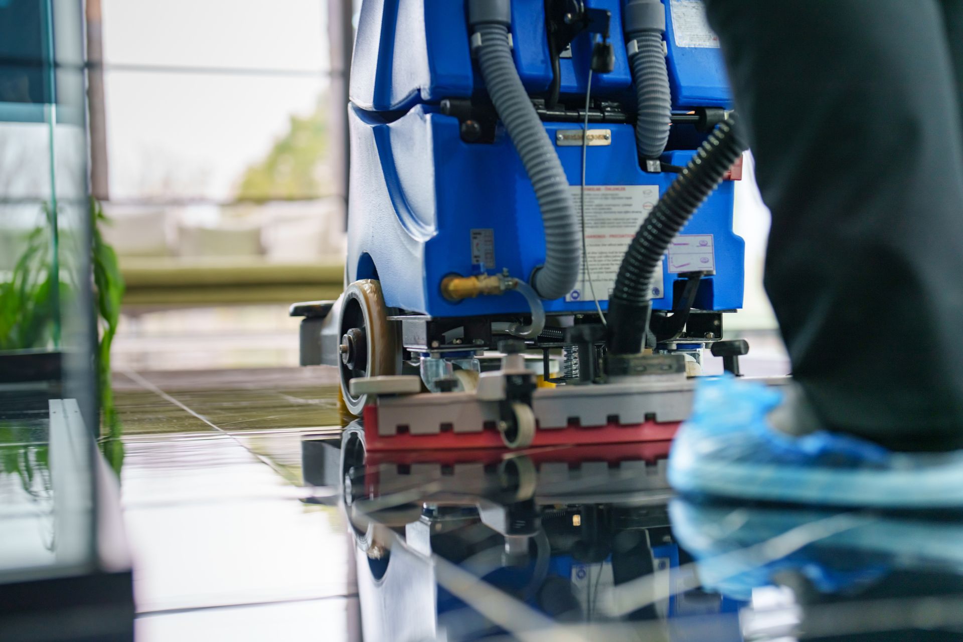 Blue floor cleaning machine in use, reflected on a shiny floor; person wearing blue shoe covers.