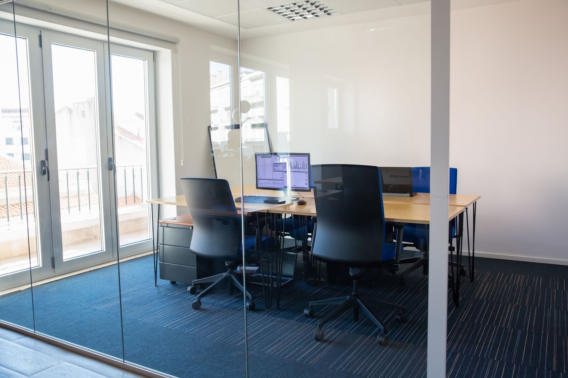 Office with desks, chairs, computer monitors. Glass wall, blue carpet, and a balcony.