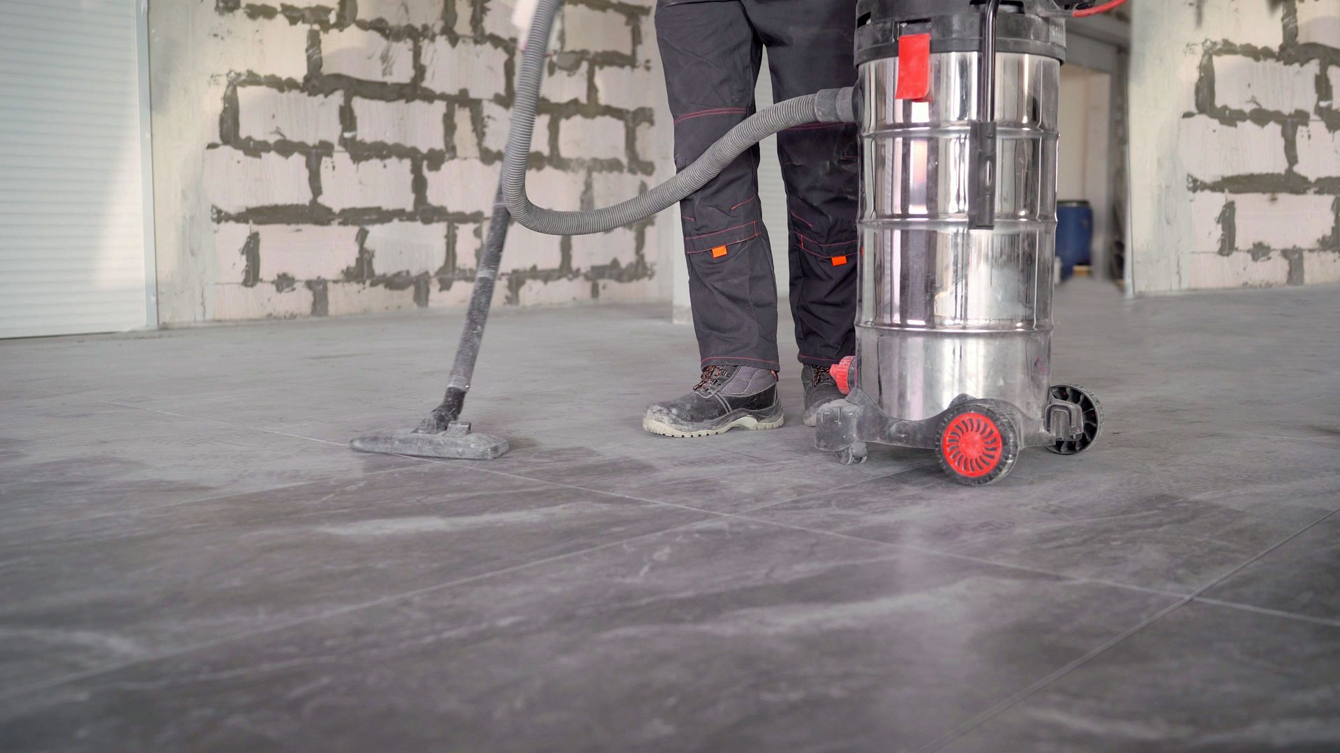 Person vacuuming a dusty concrete floor in a construction setting with a large, wheeled vacuum.