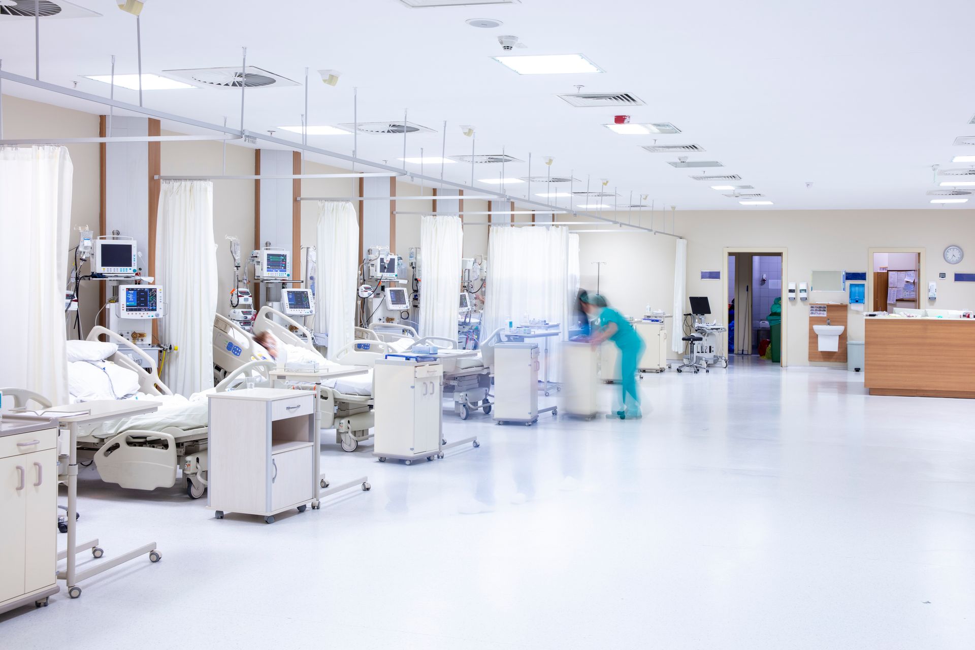Hospital room with patients in beds, medical equipment, and a blurred nurse.