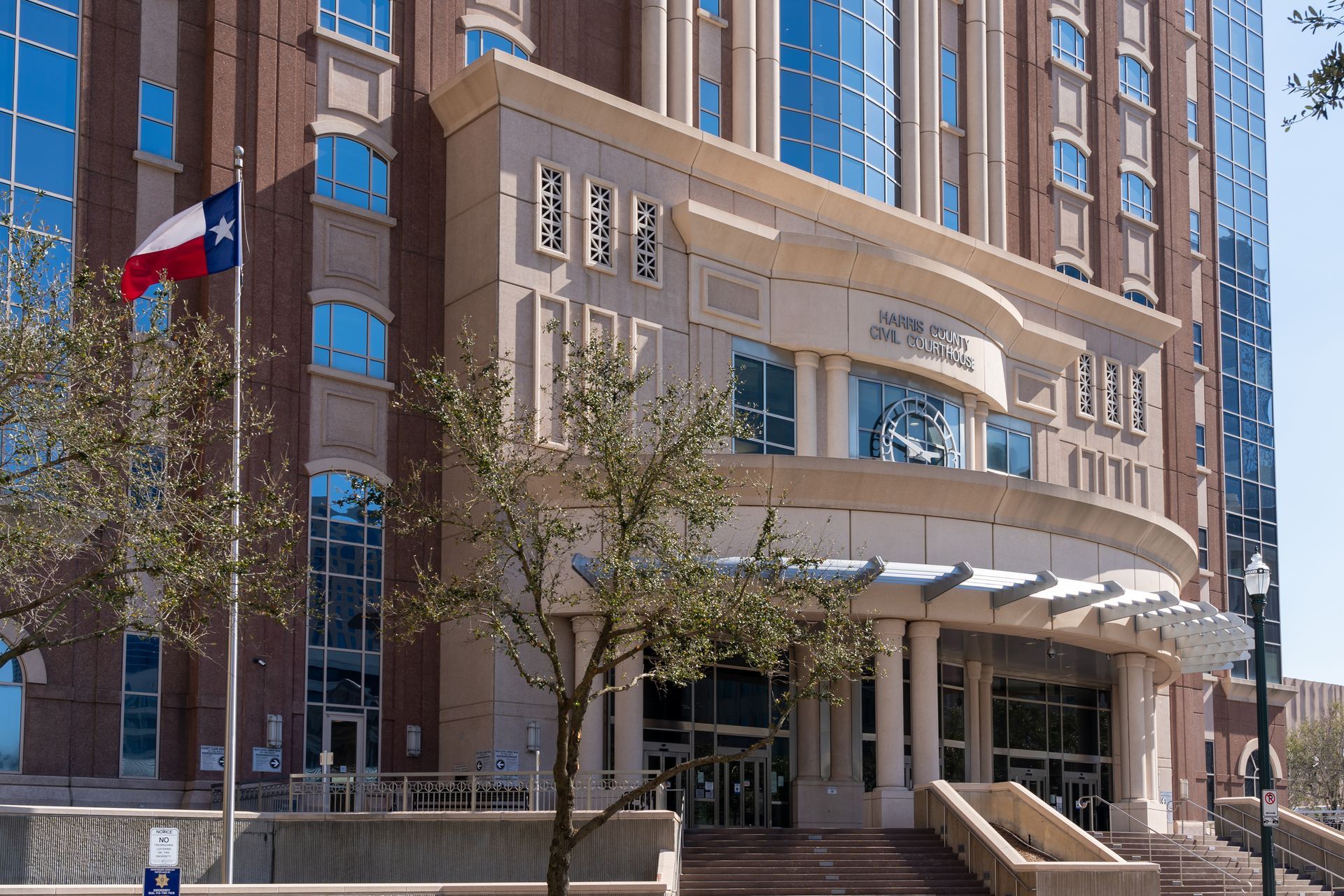 A modern courthouse with a Texas flag. Beige and brown brick building with glass windows, front entrance and tree.