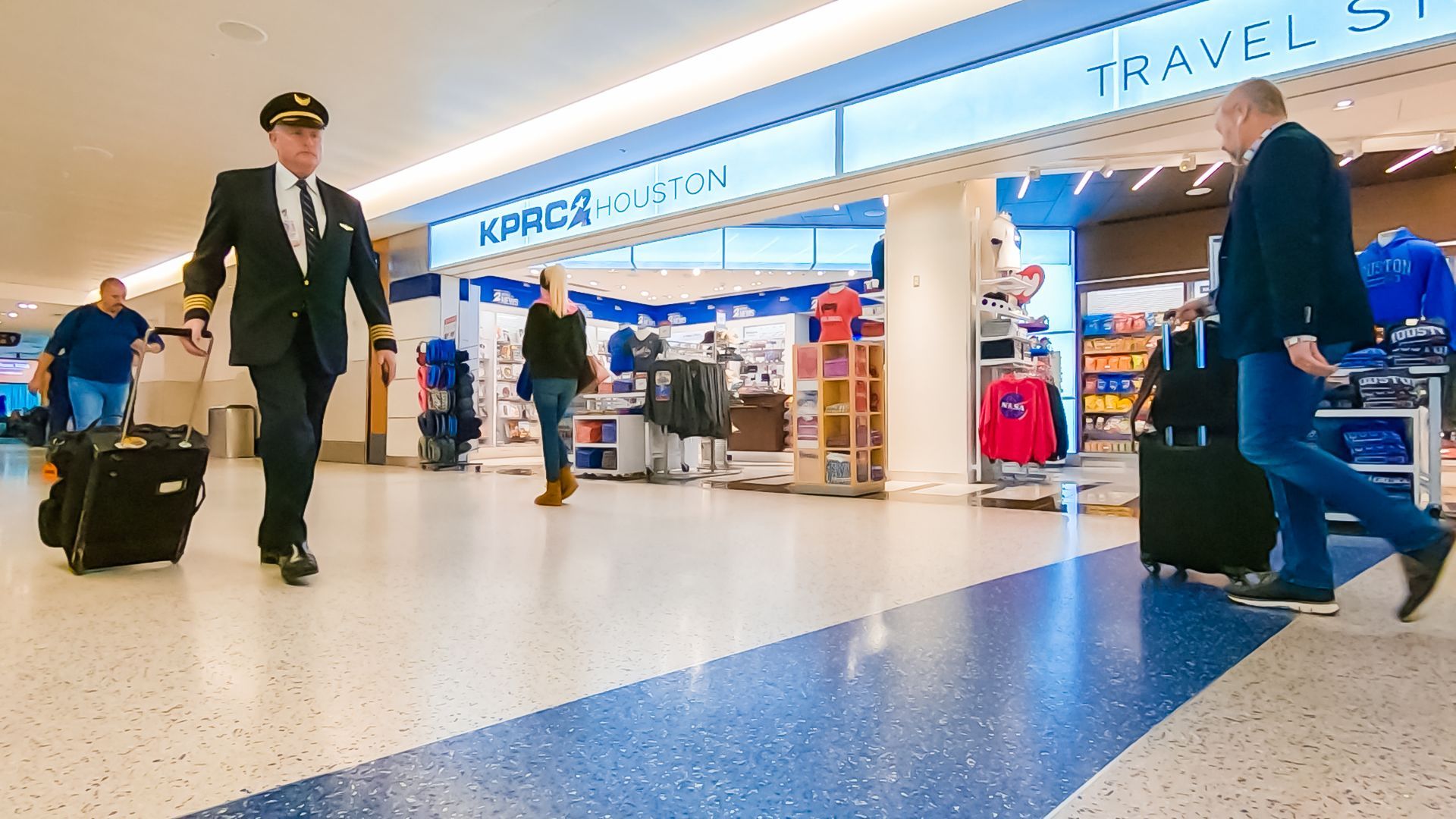 Pilot in uniform and man with luggage walk in airport terminal, near a store.