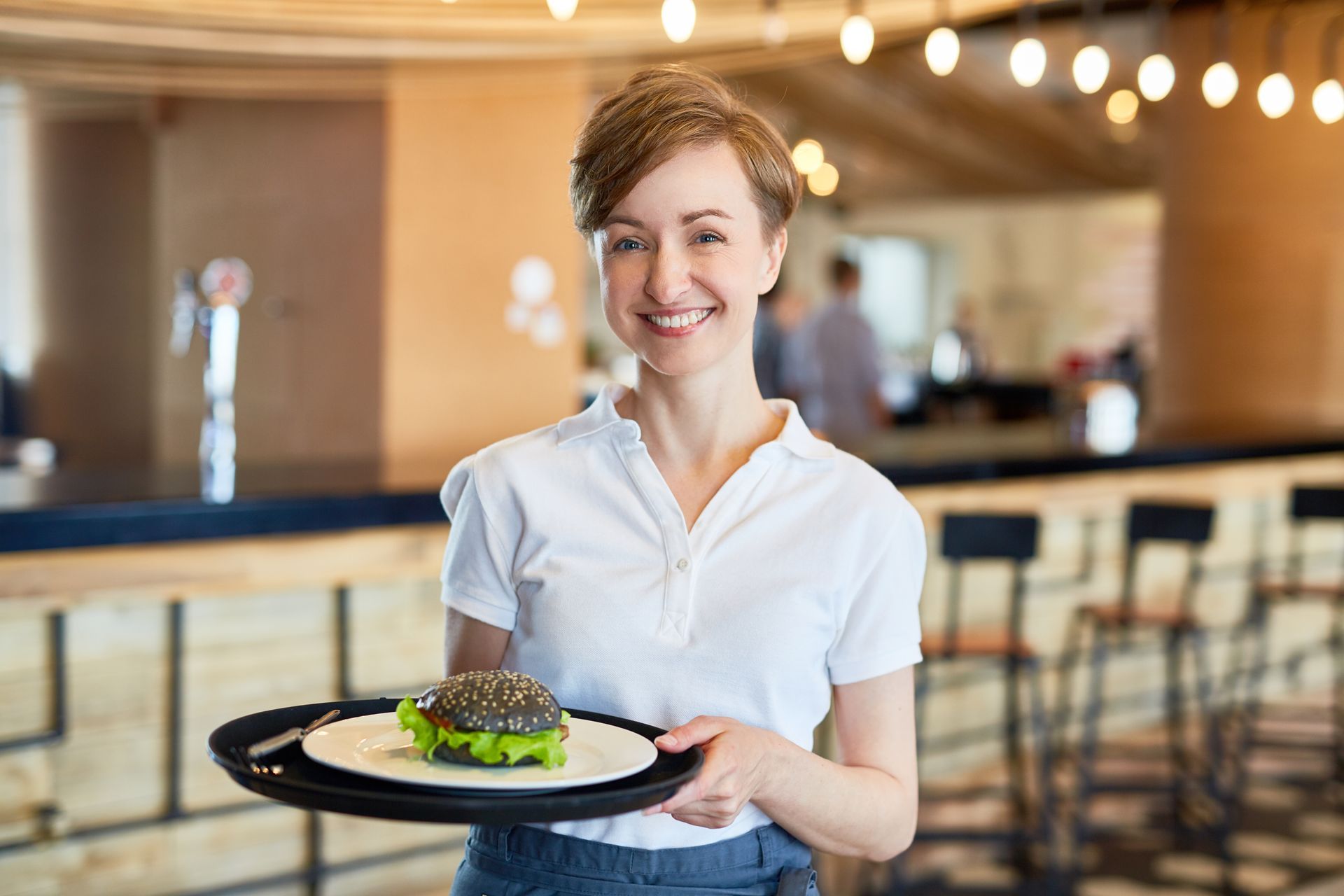 Waitress smiles, holding a tray with a burger in a restaurant.