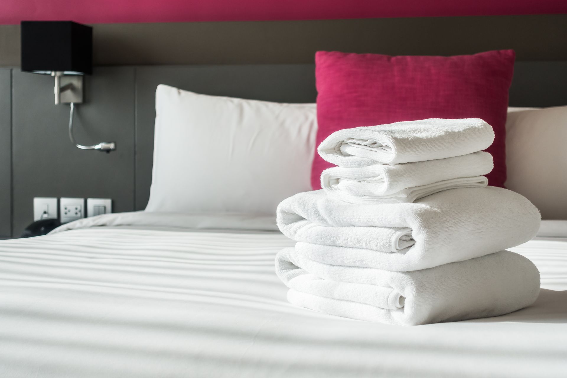 Stack of white towels on a bed with white and pink pillows; a nightstand lamp is above the bed.