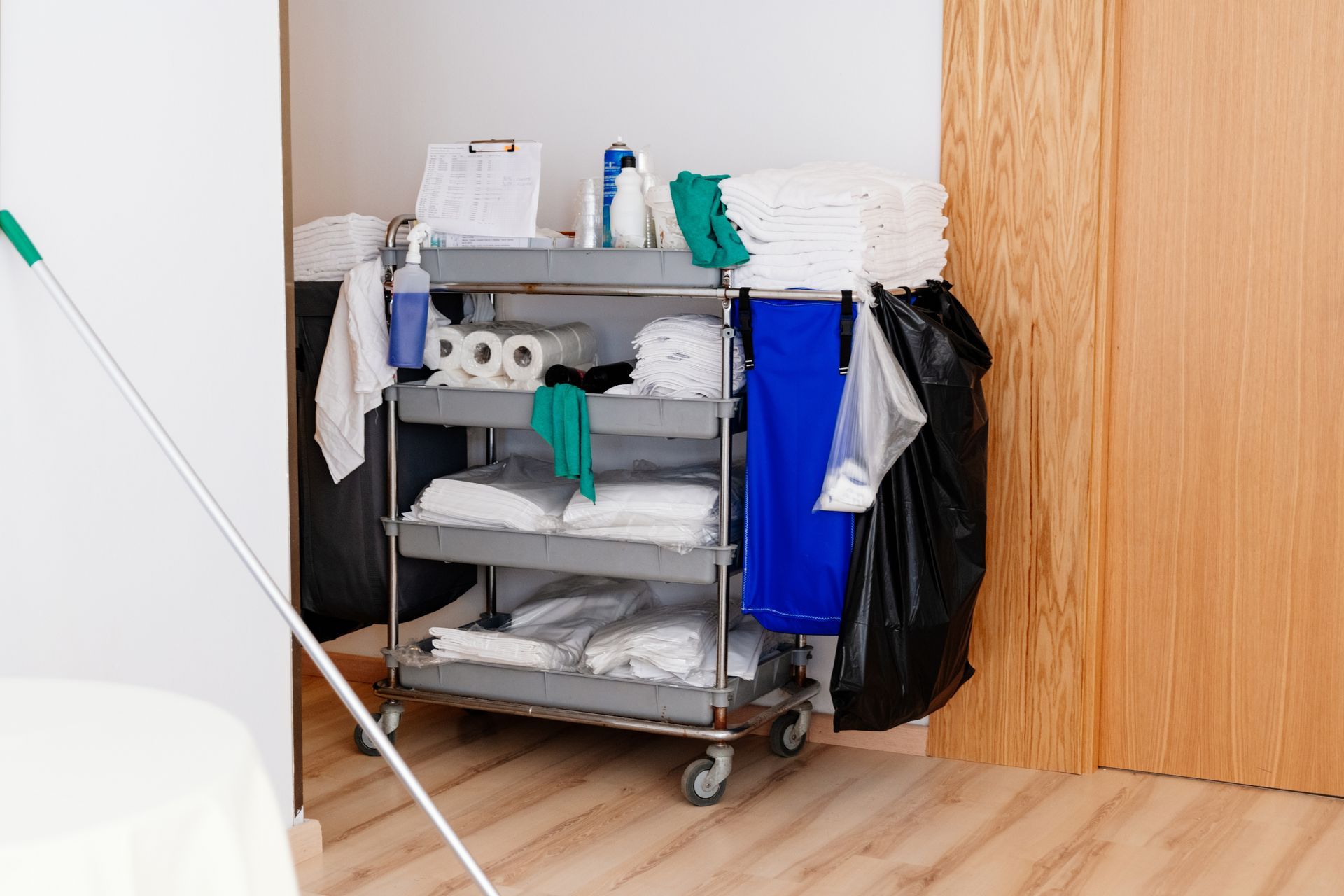 Housekeeping cart with cleaning supplies, towels, and trash bag, near a wall and door.