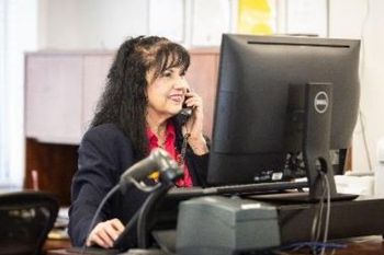 Woman in blazer on phone at desk, looking at a computer screen.