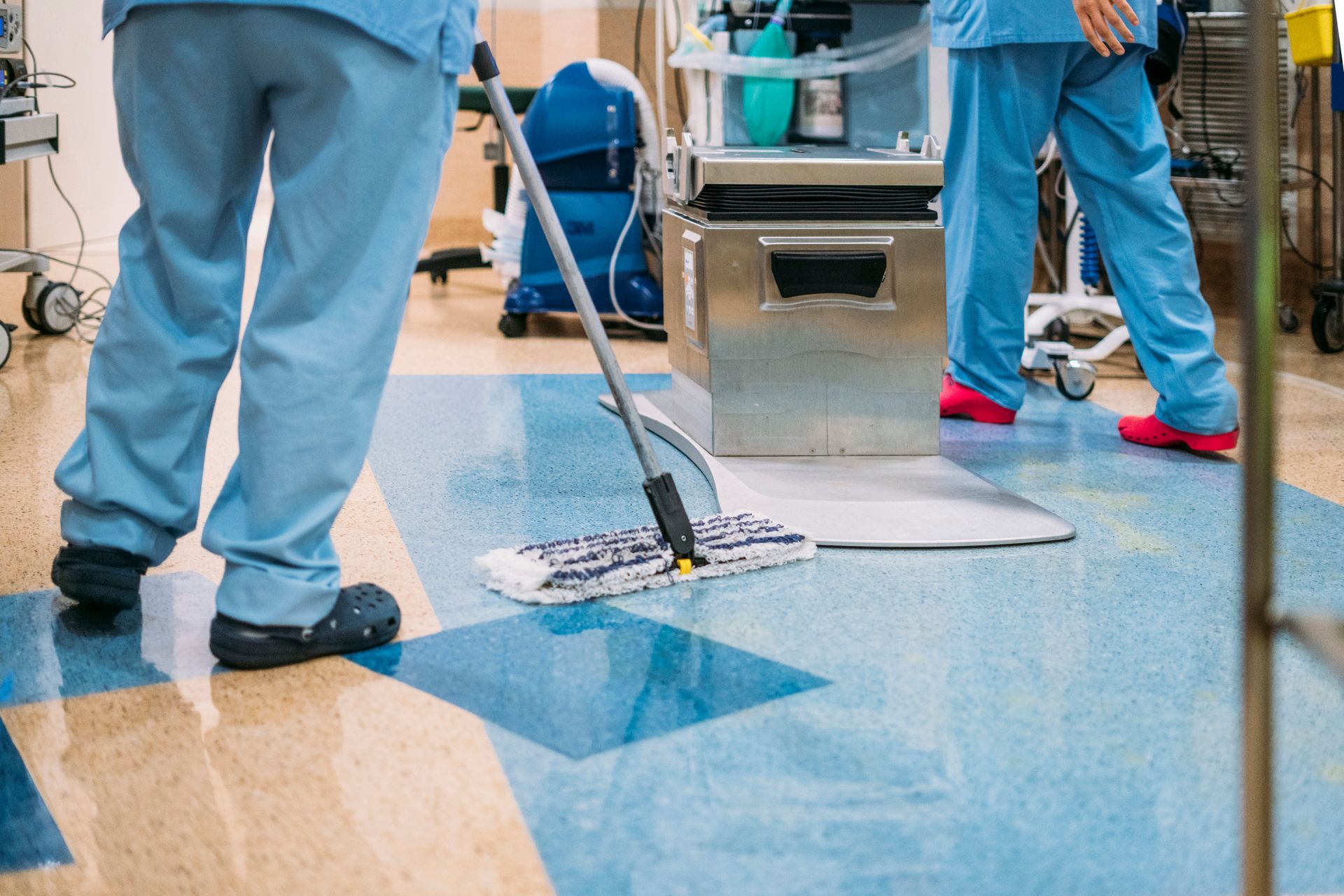 Two figures in blue scrubs mopping a floor in a medical setting, with equipment visible.