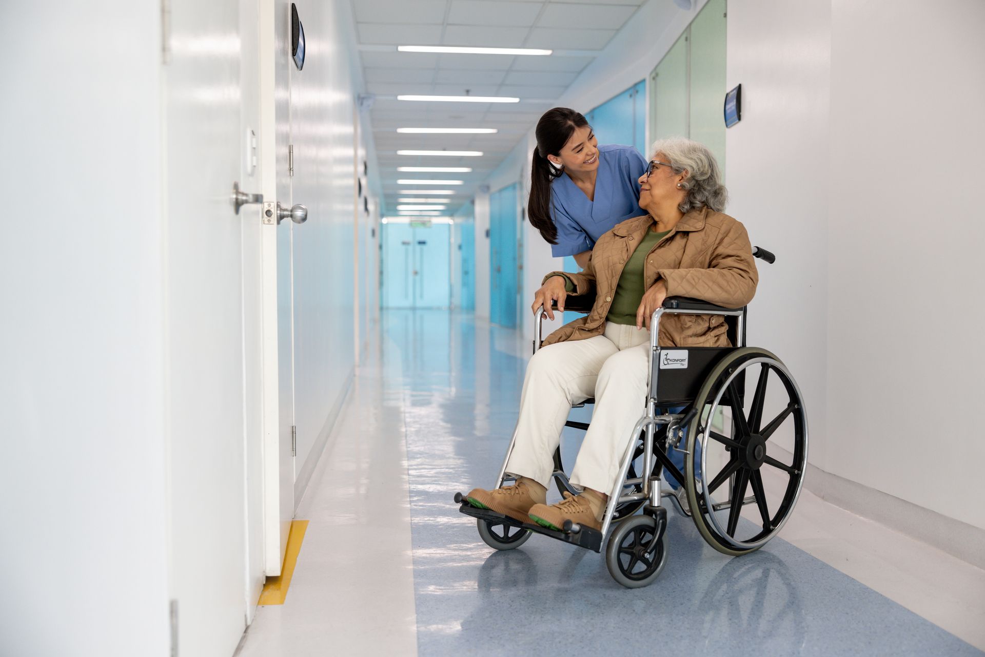 Nurse assists elderly person in wheelchair down a hospital hallway.