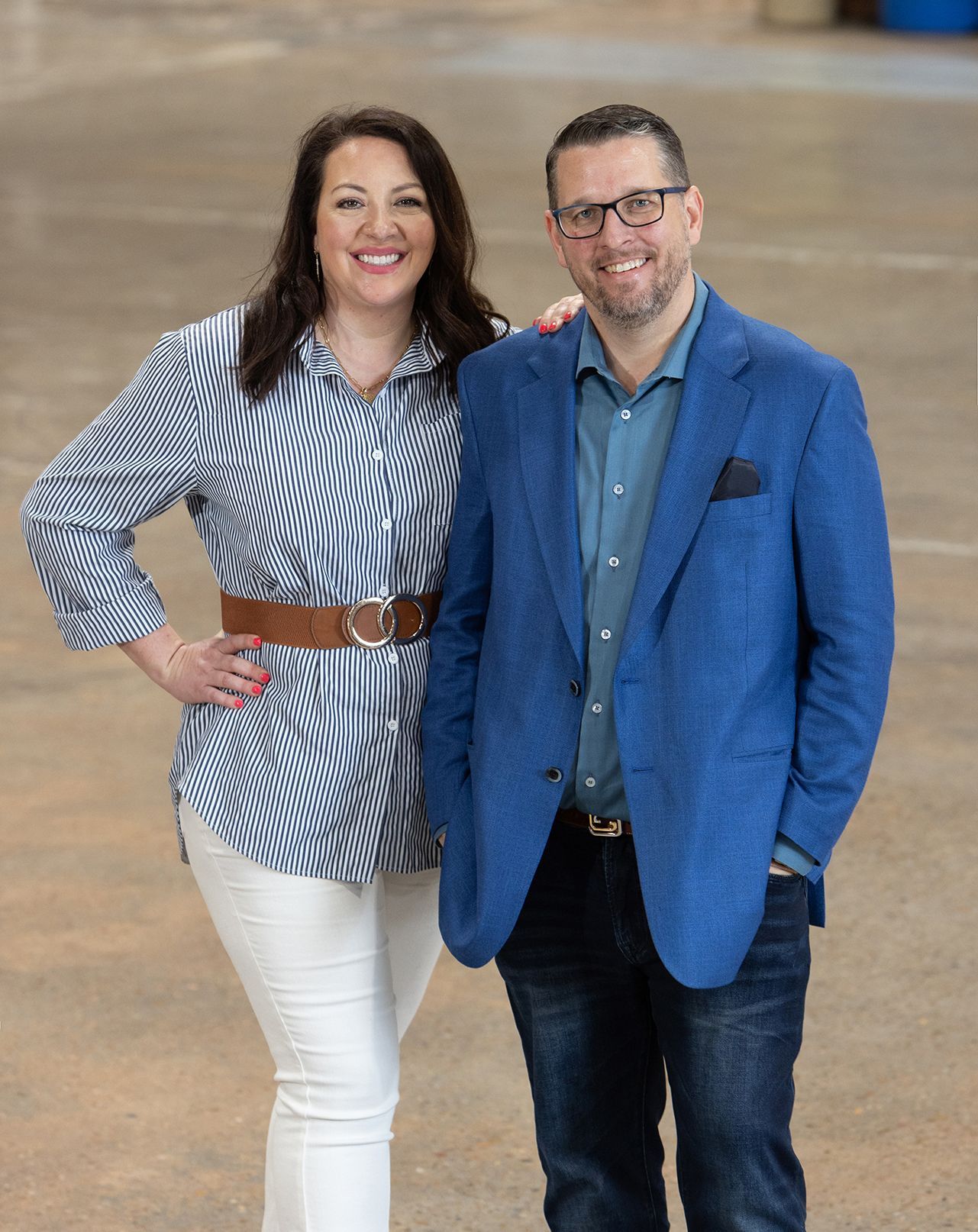 Two people standing indoors, smiling, wearing a blue blazer and striped shirt with white pants.