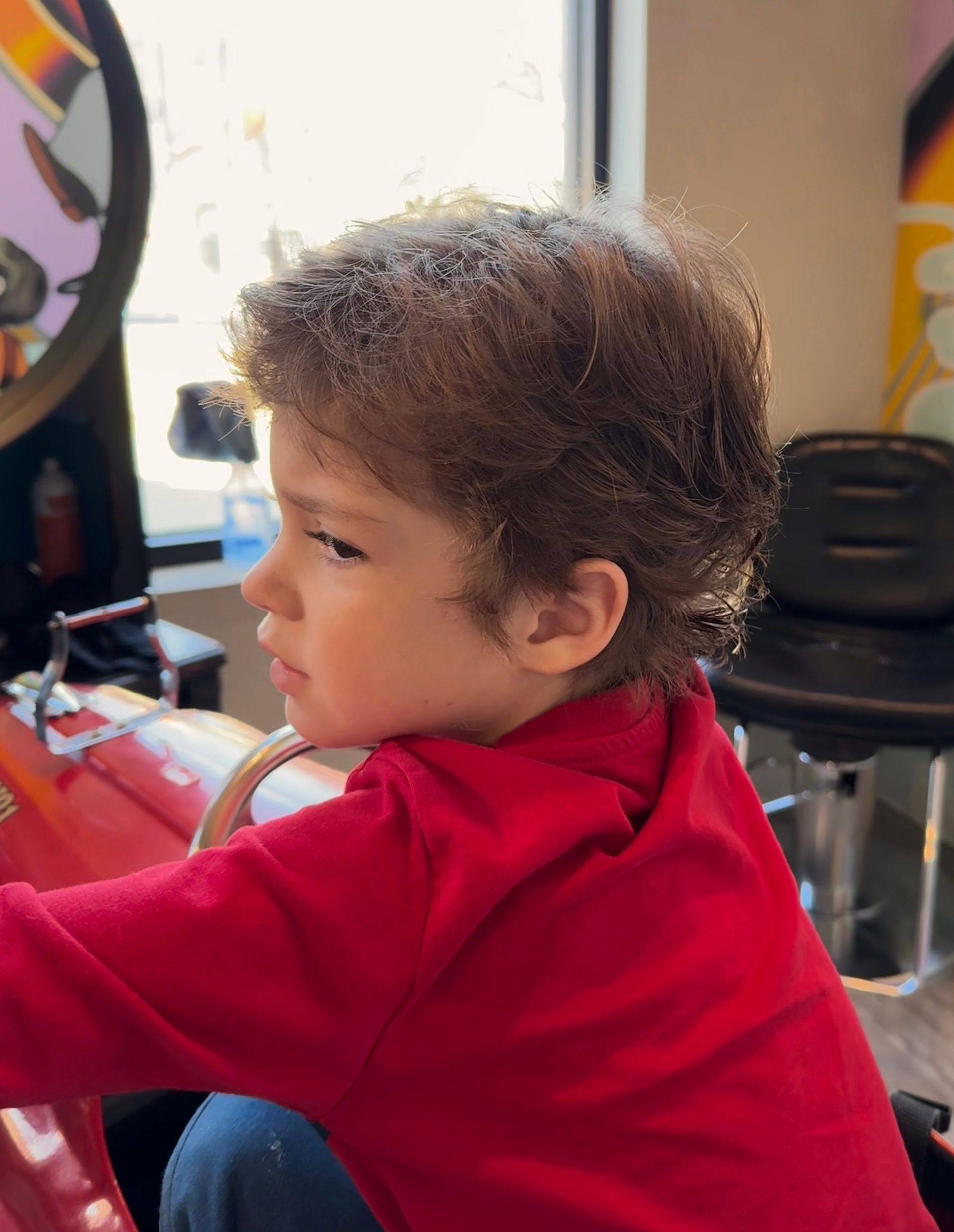 Toddler in a red shirt seated indoors, looking left beside a table and chair.
