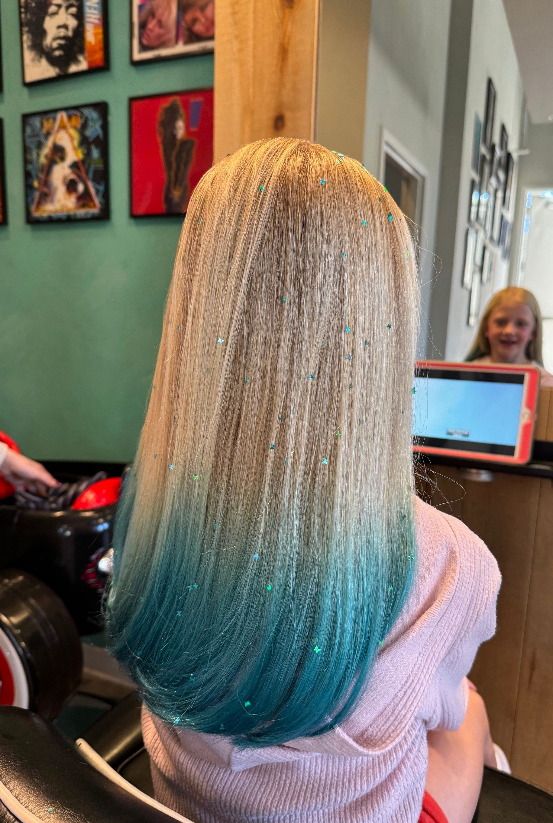 Long blonde-to-blue ombré hair seen from behind in a salon chair, with a stylist in the background