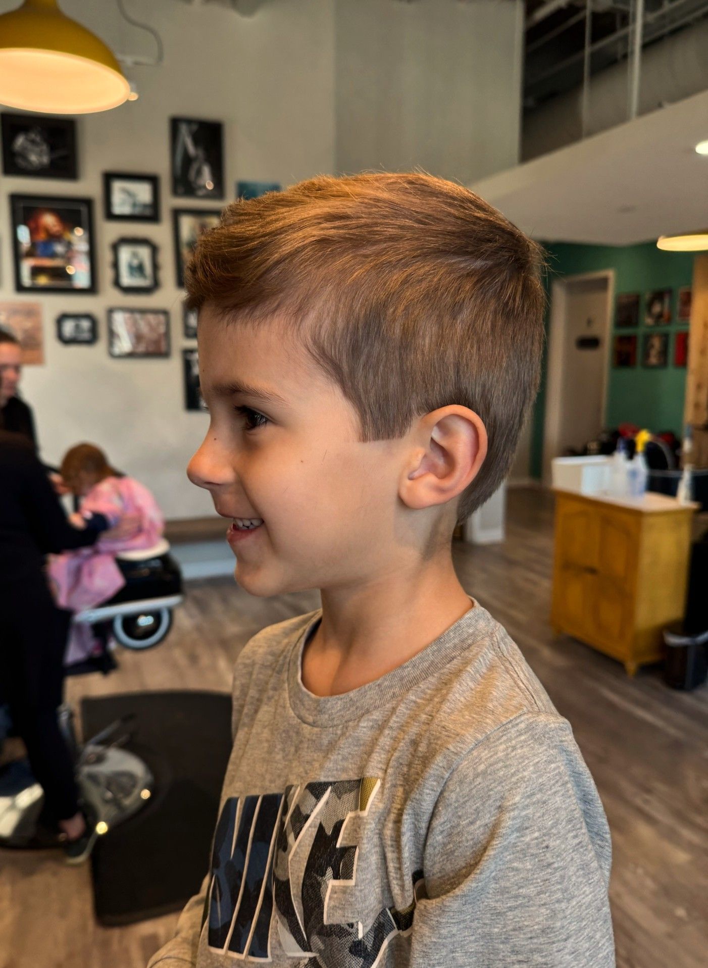 Boy smiling in a barber shop, wearing a gray Nike shirt, with salon chairs and wall photos in the background