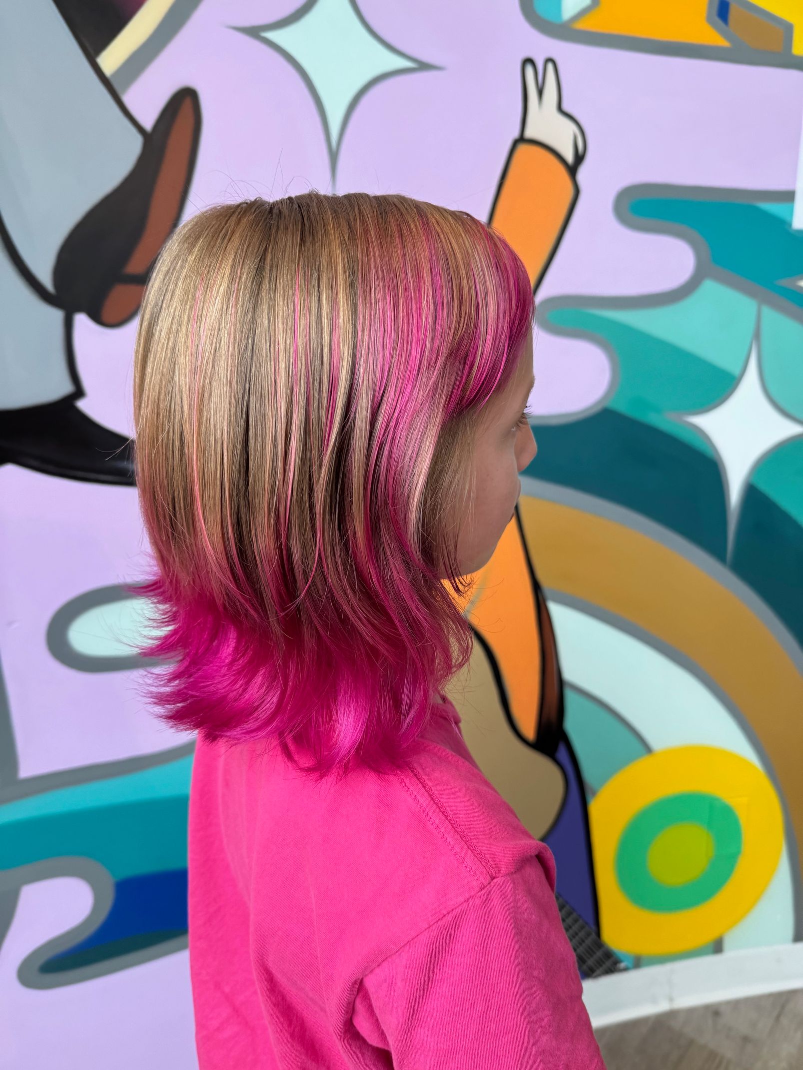 Side view of a child with pink-dyed hair in front of a colorful graffiti mural