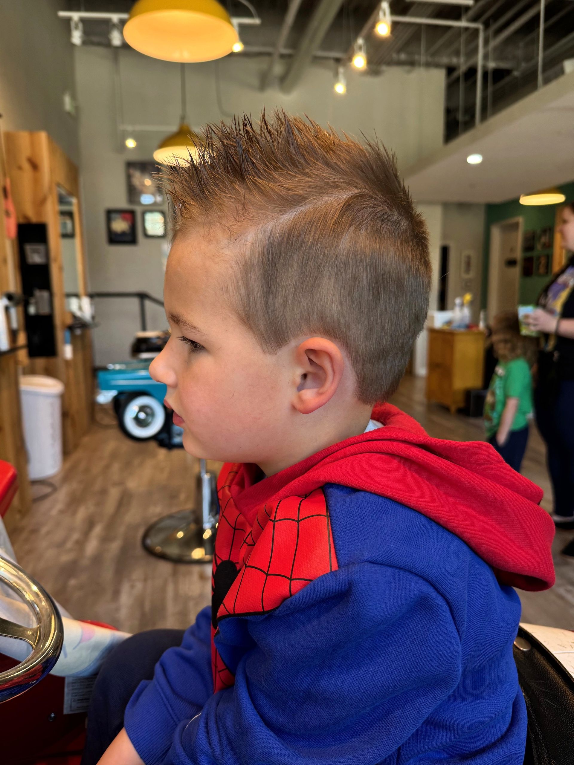 Child in a red-and-blue hoodie seated with new hair style.