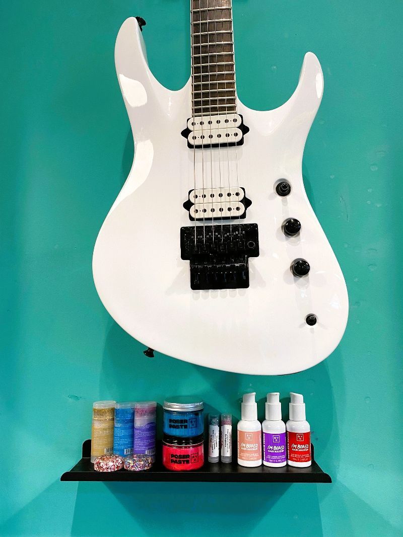 A white electric guitar is hanging on a blue wall next to a shelf with nail polish.