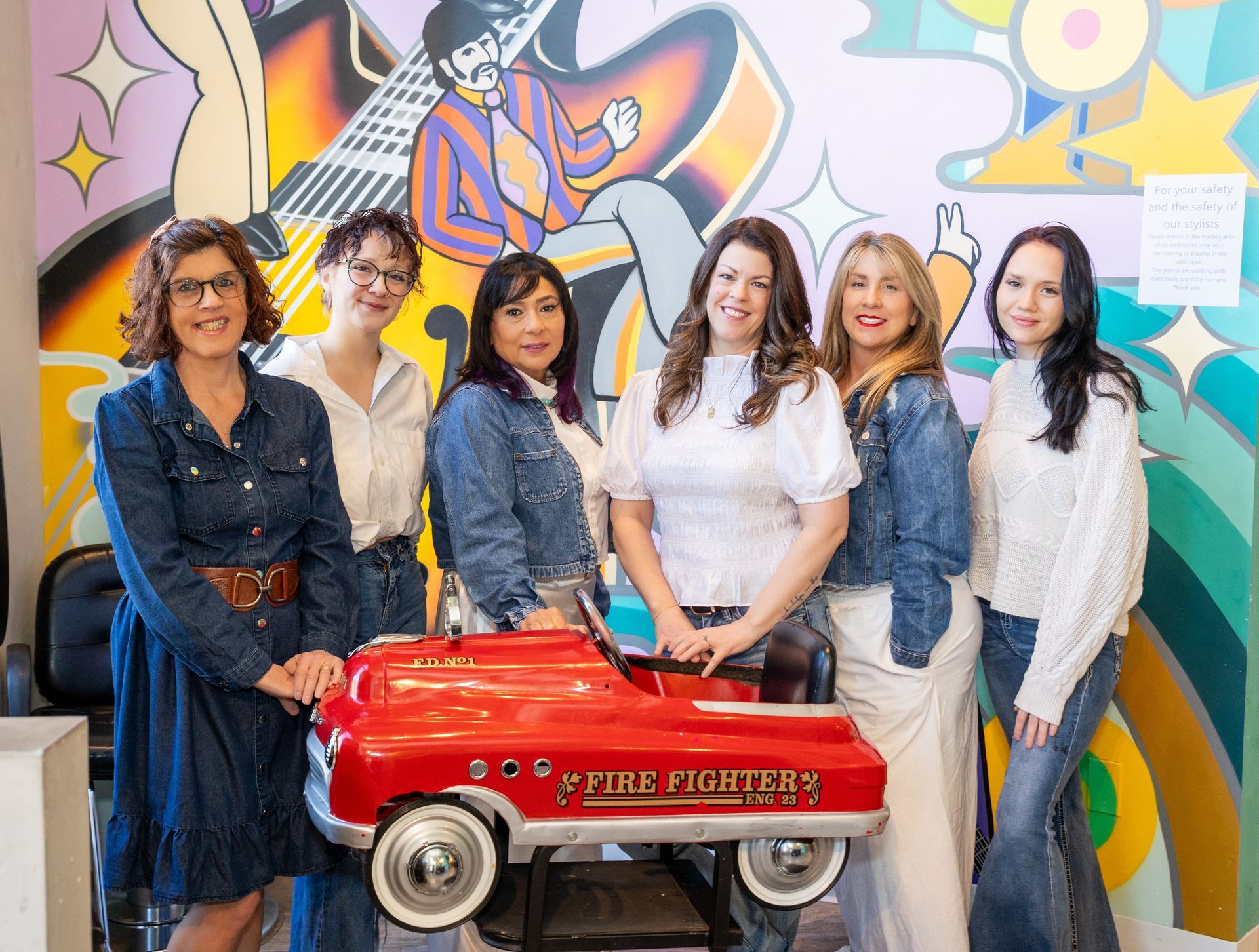 Six women pose with a red fire truck toy in front of a colorful mural.