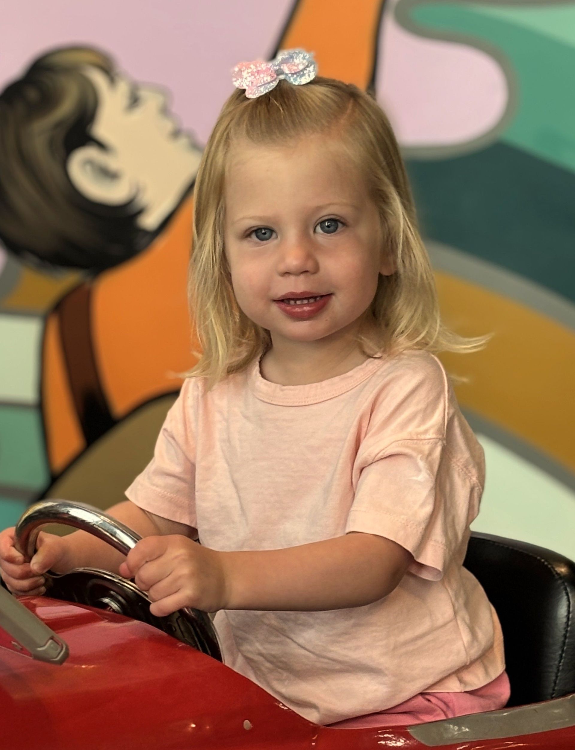 A little girl in a pink shirt is sitting in a red toy car.