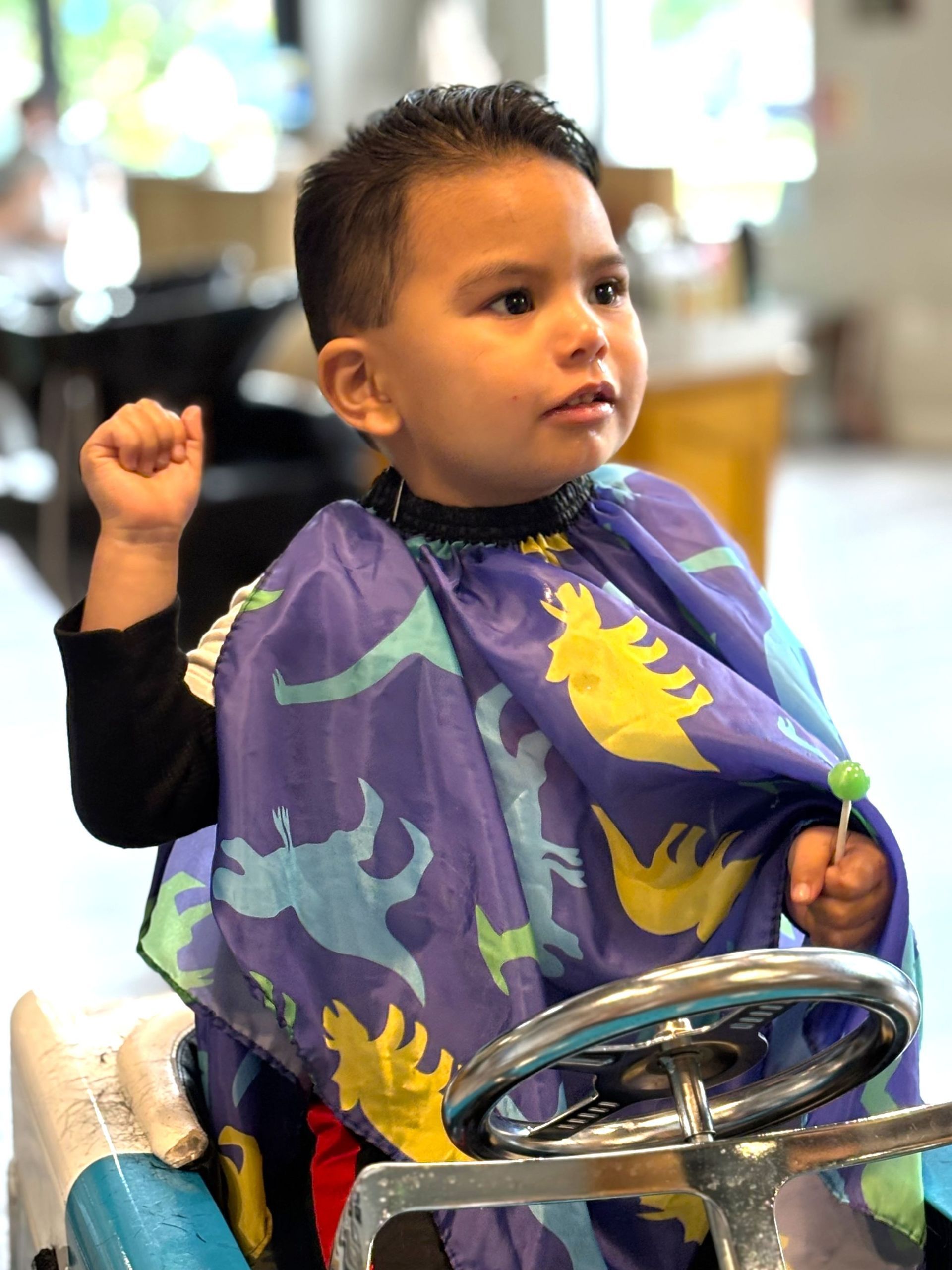 A young boy is sitting in a barber chair wearing a purple cape with dinosaurs on it.