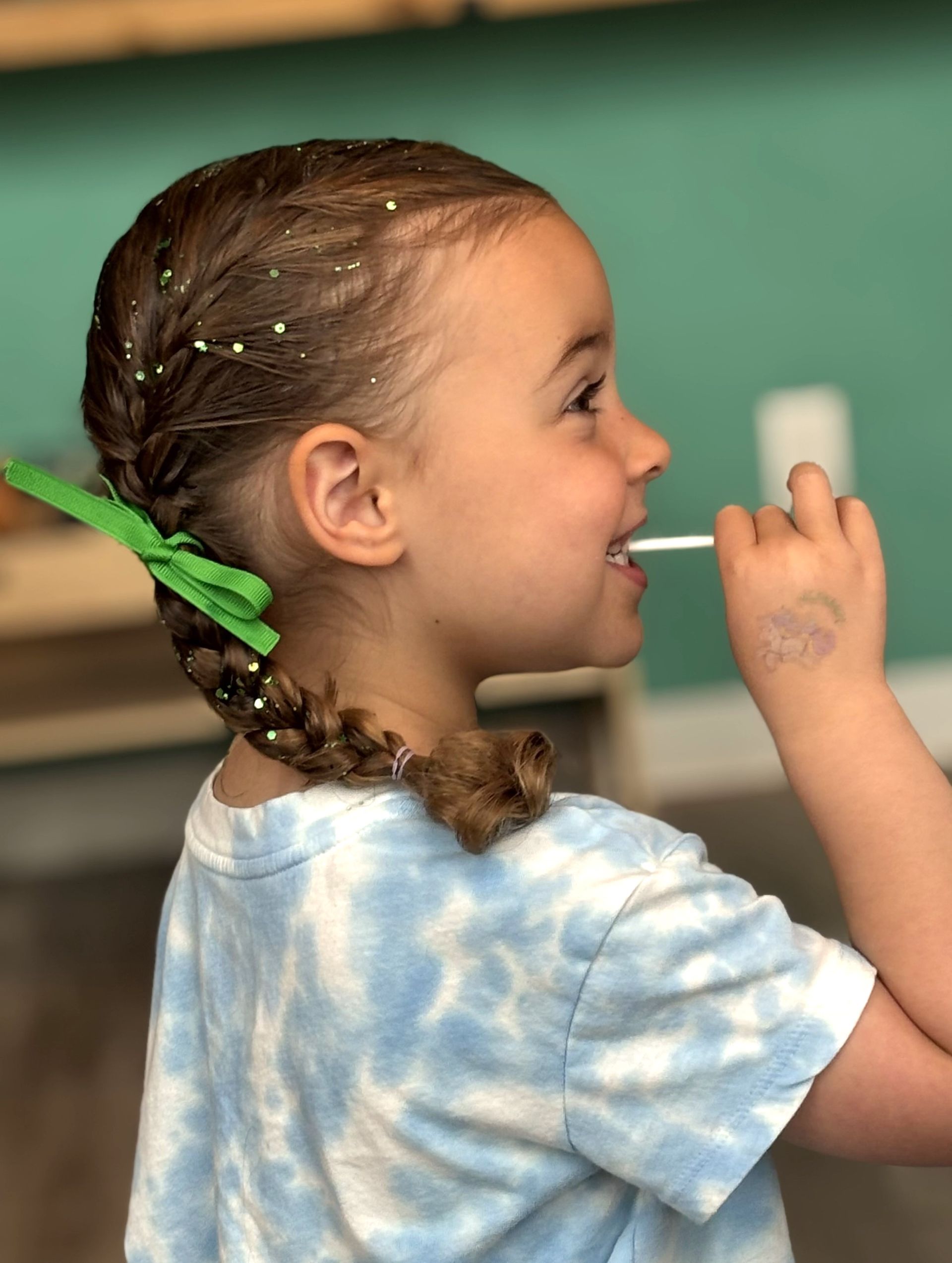 A little girl in a blue tie dye shirt is eating a lollipop.