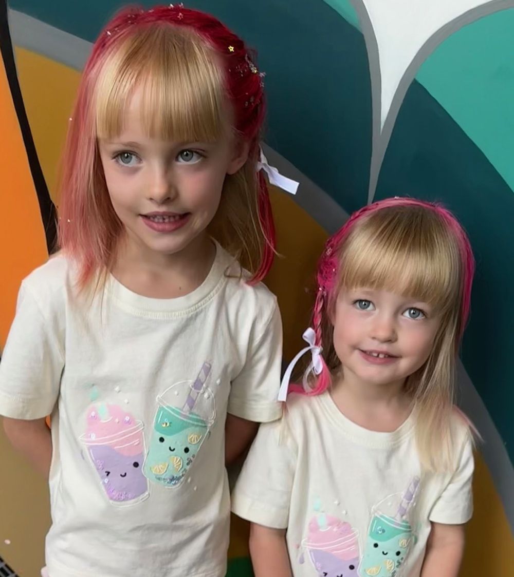 Two young girls with blonde hair and matching t-shirts posing in front of a colorful wall.