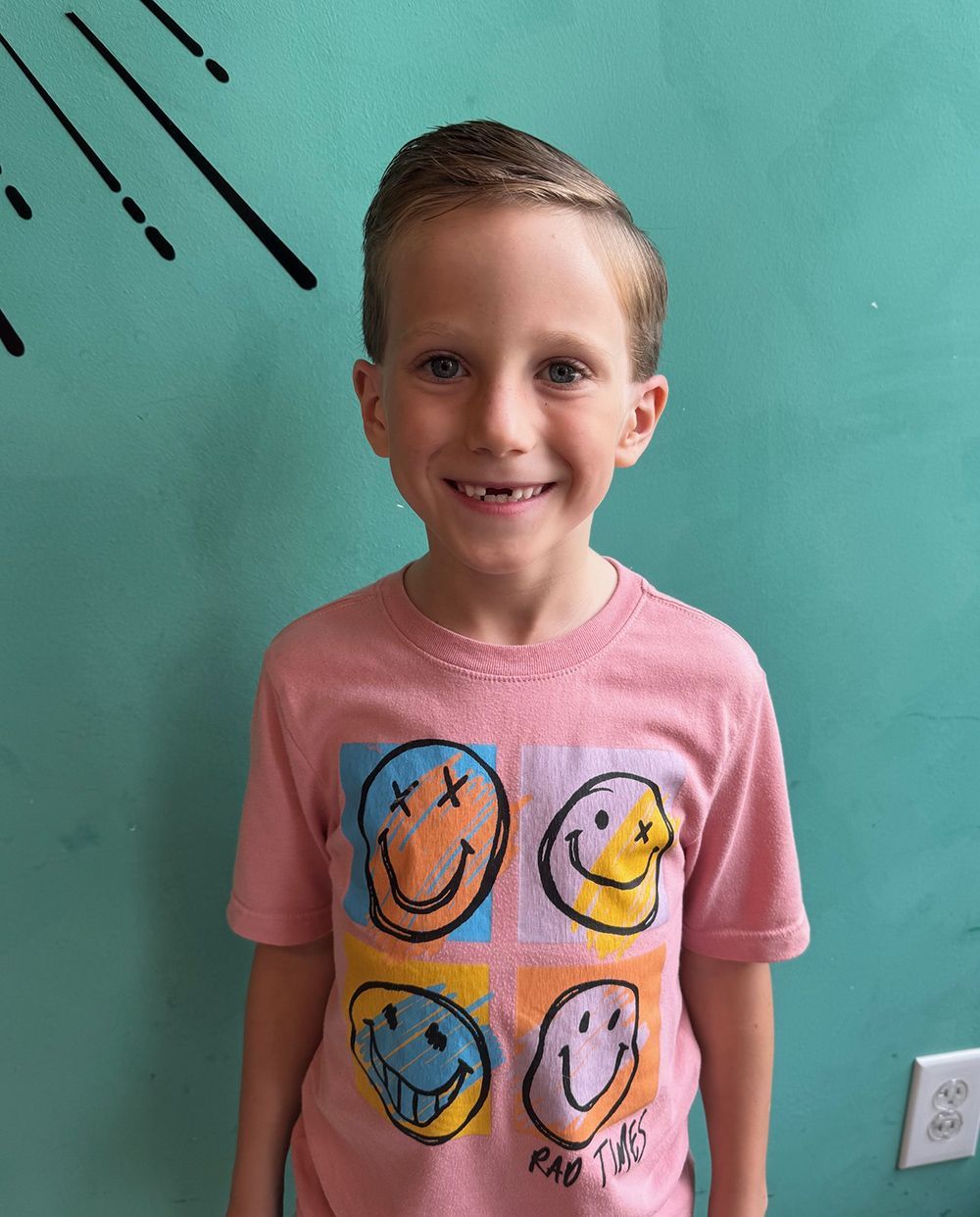 Young boy wearing a pink shirt with smiley faces, in front of a teal wall.