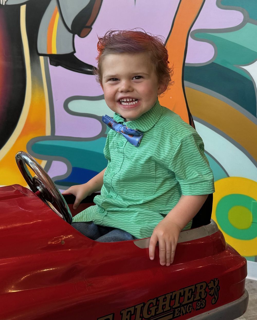 Boy with red curly hair, smiles at the camera while driving a red toy truck.
