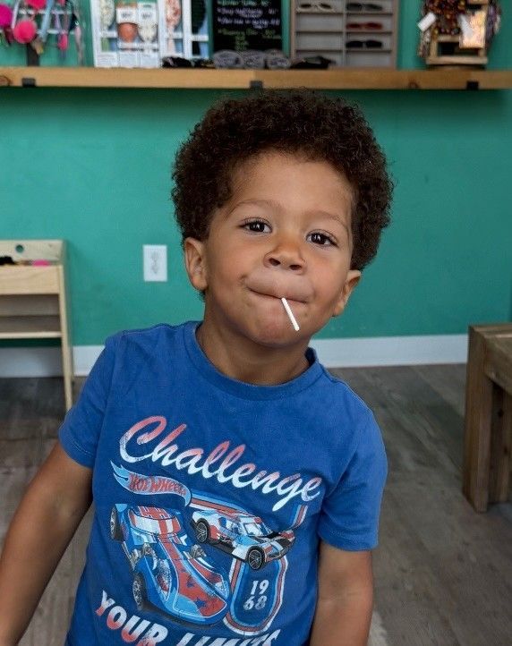 Young boy with curly hair, lollipop in mouth, wearing a blue t-shirt.