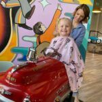 A little girl is sitting in a toy car getting her hair cut.