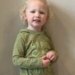 A little girl is standing in front of a wall wearing a green shirt.