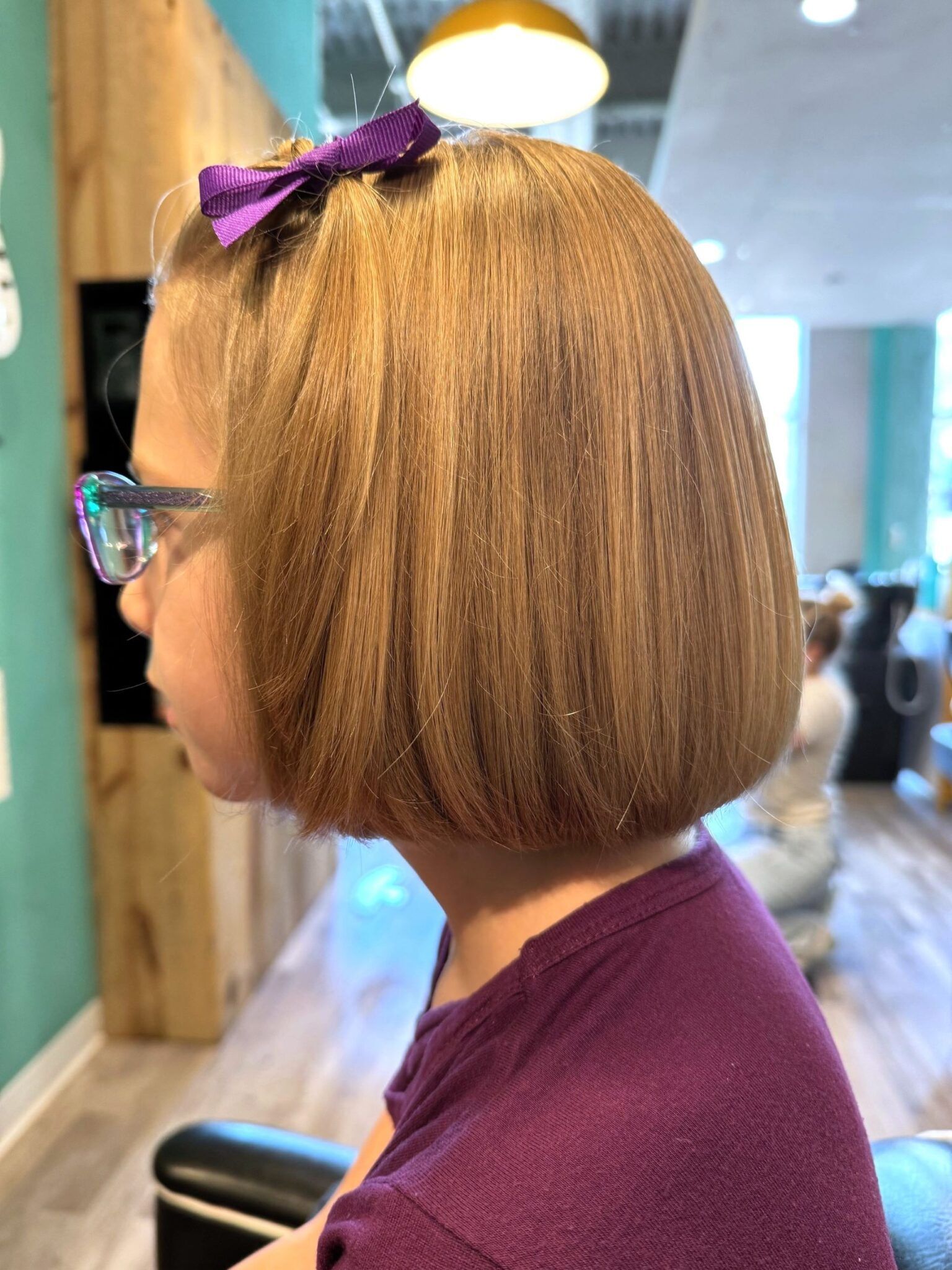 A little girl with glasses and a bow in her hair is sitting in a chair.