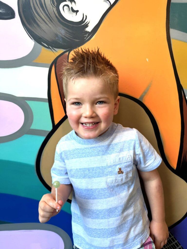 A young boy is holding a lollipop in front of a colorful wall.