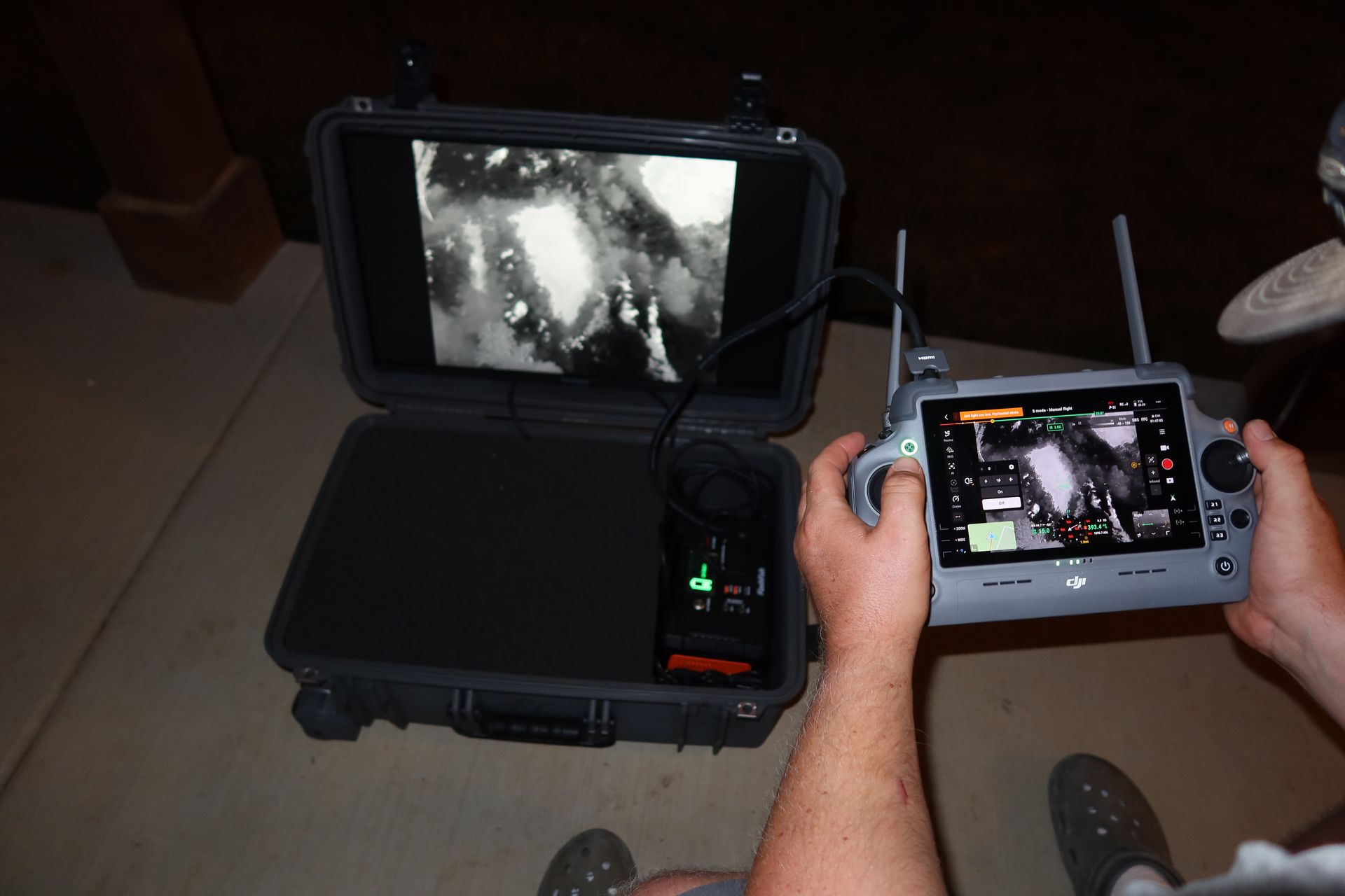 Hands using a handheld controller and tablet to view a live camera feed from a case-mounted screen.